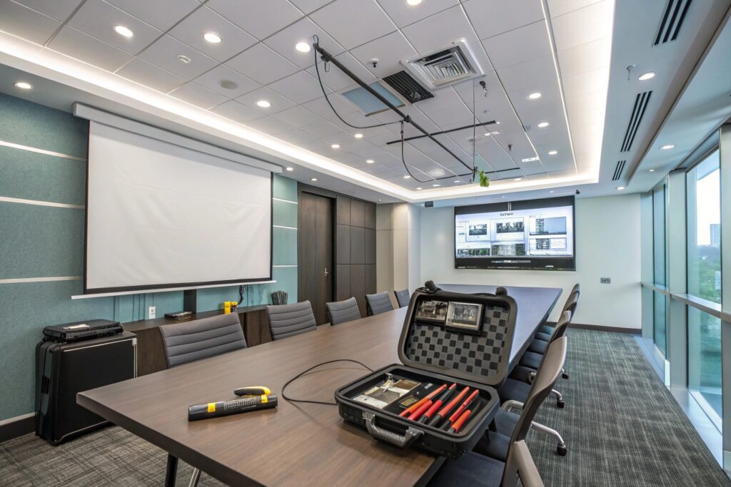 Sleek conference room with a large wooden table, black ergonomic chairs, advanced tech equipment, open tool case, projection screen, and digital display, teal and gray color scheme, large windows, recessed lighting, patterned carpet, ideal for professional business presentations.